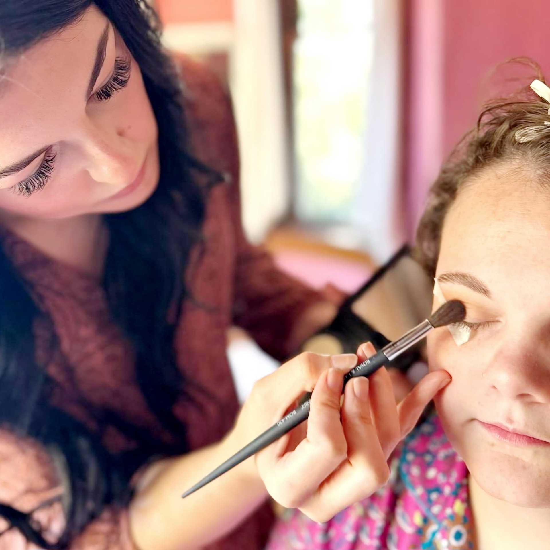 Makeup artist applying eyeshadow to a woman with closed eyes in a softly lit room.
